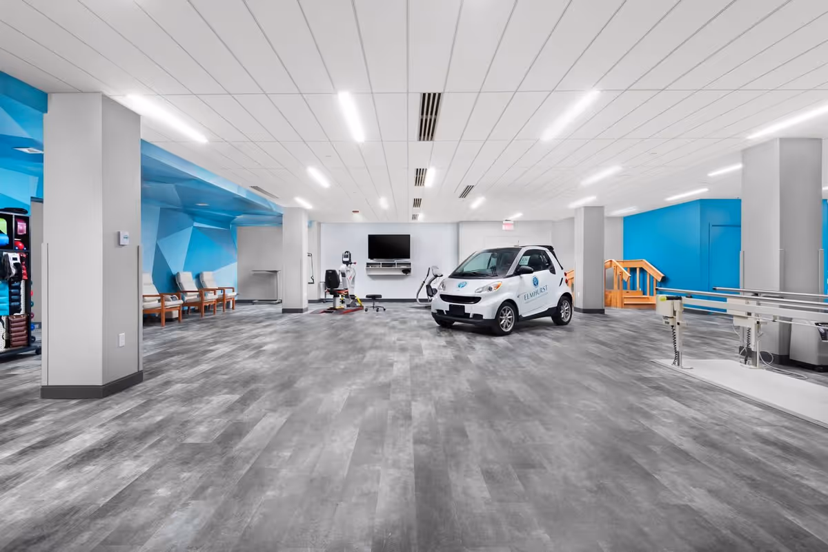 A spacious rehabilitation room with gray flooring and white ceiling lights. The room features a small white car with Elmhurst logo, exercise equipment, a wall-mounted TV, chairs along a blue geometric accent wall, and parallel bars for physical therapy. There is a wooden staircase leading to a blue wall area in the background.