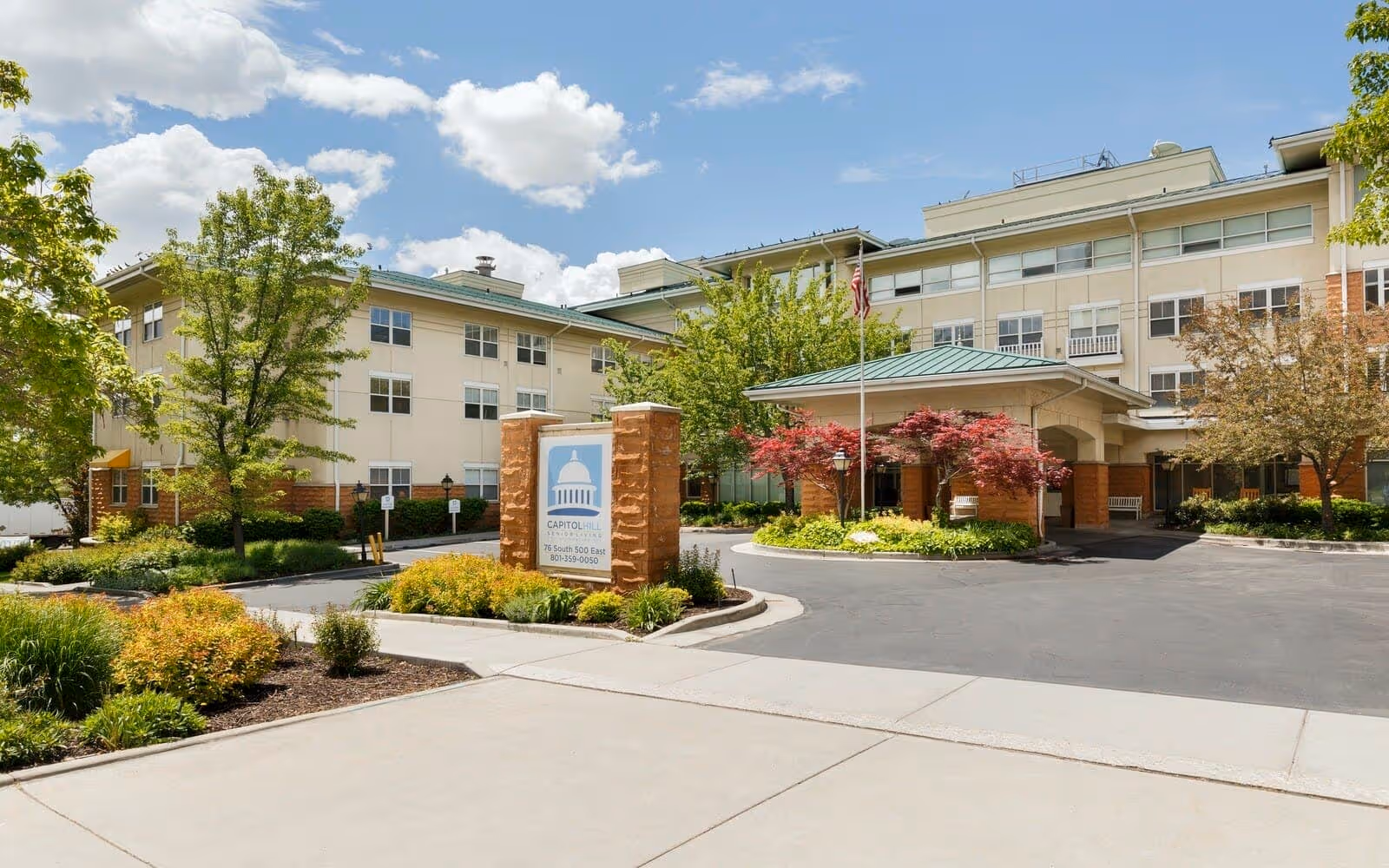 Exterior view of Capitol Hill Assisted Living & Memory Care facility showing a multi-story building with beige walls and green roof. There is a covered entrance with an American flag and landscaped greenery including trees and bushes around the driveway and sidewalk.