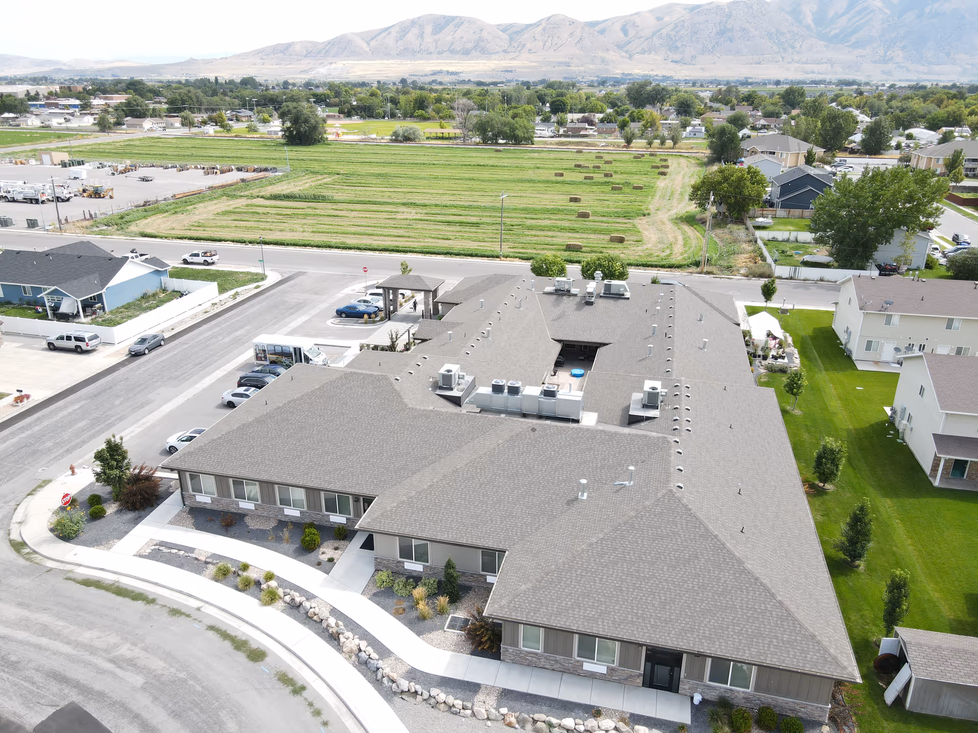 Aerial view of a single-story assisted living facility building with a gray roof, surrounded by parking spaces, green lawns, and nearby residential houses. In the background, there are open fields and mountains under a clear sky.