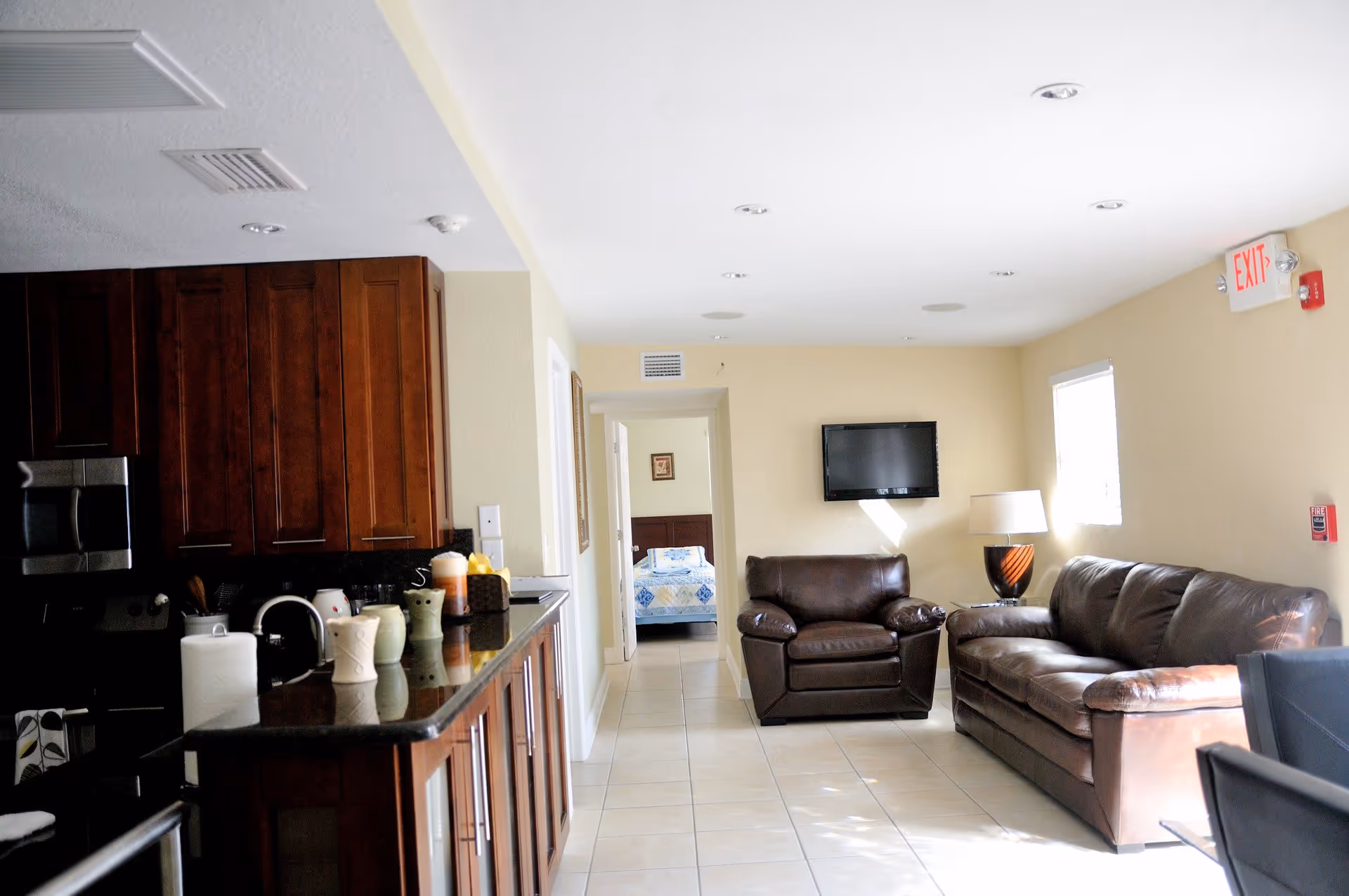Interior view of a senior living facility showing a living room area with a brown leather sofa and armchair, a wall-mounted TV, a lamp on a side table, and a kitchen area with dark wood cabinets and a black countertop. A bedroom with a bed covered in a blue and white quilt is visible through an open doorway.