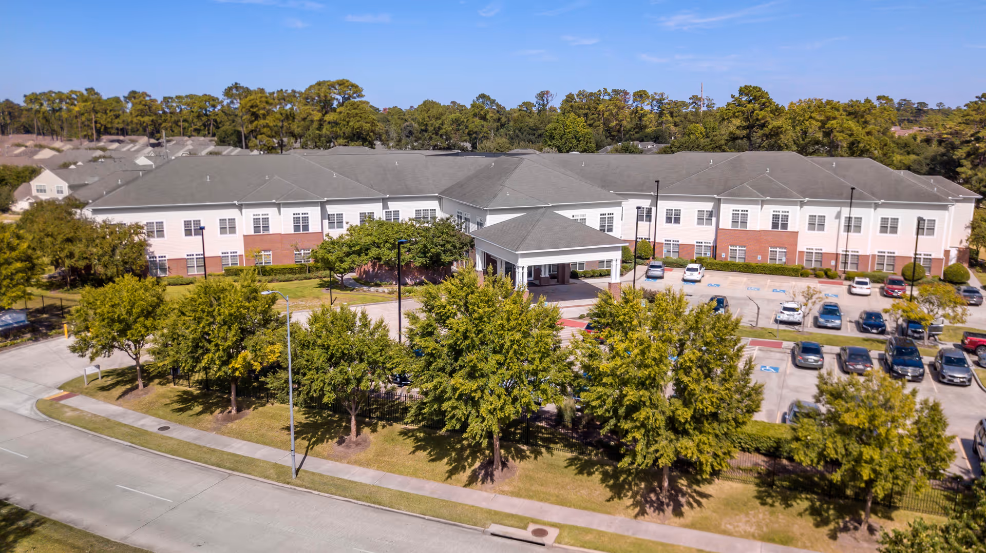 Aerial view of a large two-story senior living facility building with a covered entrance, surrounded by trees and a parking lot with several cars. The building has a combination of white siding and red brick exterior walls, with a gray roof. The area is landscaped with green grass and trees, and a clear blue sky is visible above.