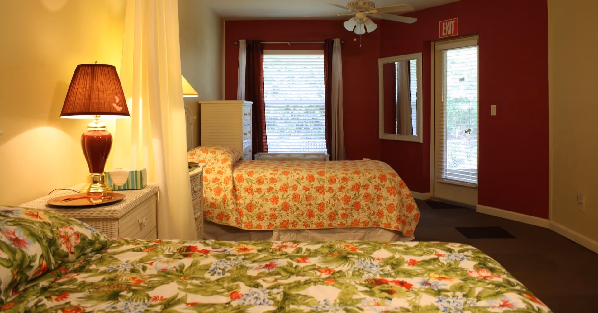 Interior view of a bedroom in an assisted living facility featuring two beds with floral bedspreads, a bedside table with a lamp, a window with blinds and curtains, a mirror on the wall, and a door with an exit sign above it.