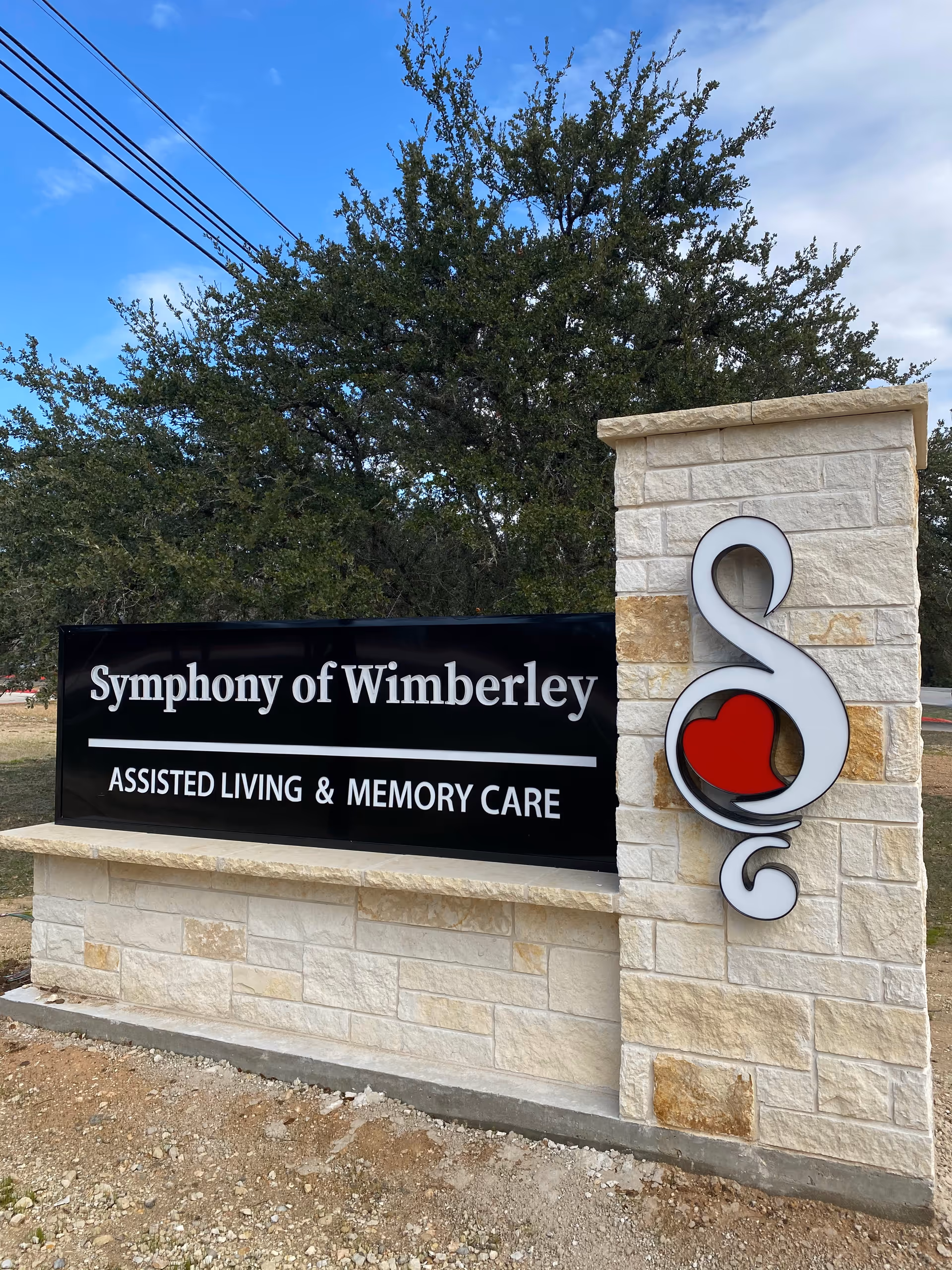 Outdoor stone sign for Symphony of Wimberley assisted living and memory care facility with a decorative logo featuring a red heart on the right side, set against a background of trees and a partly cloudy sky.