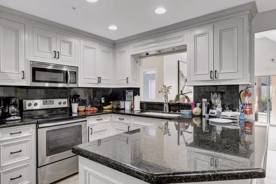 Modern kitchen with white cabinetry, black granite countertops, stainless steel oven and microwave, coffee maker, blender, and various kitchen items on the counter. There is a pass-through window with a view into another room and a fire extinguisher mounted on the wall near the entrance to an outdoor area.