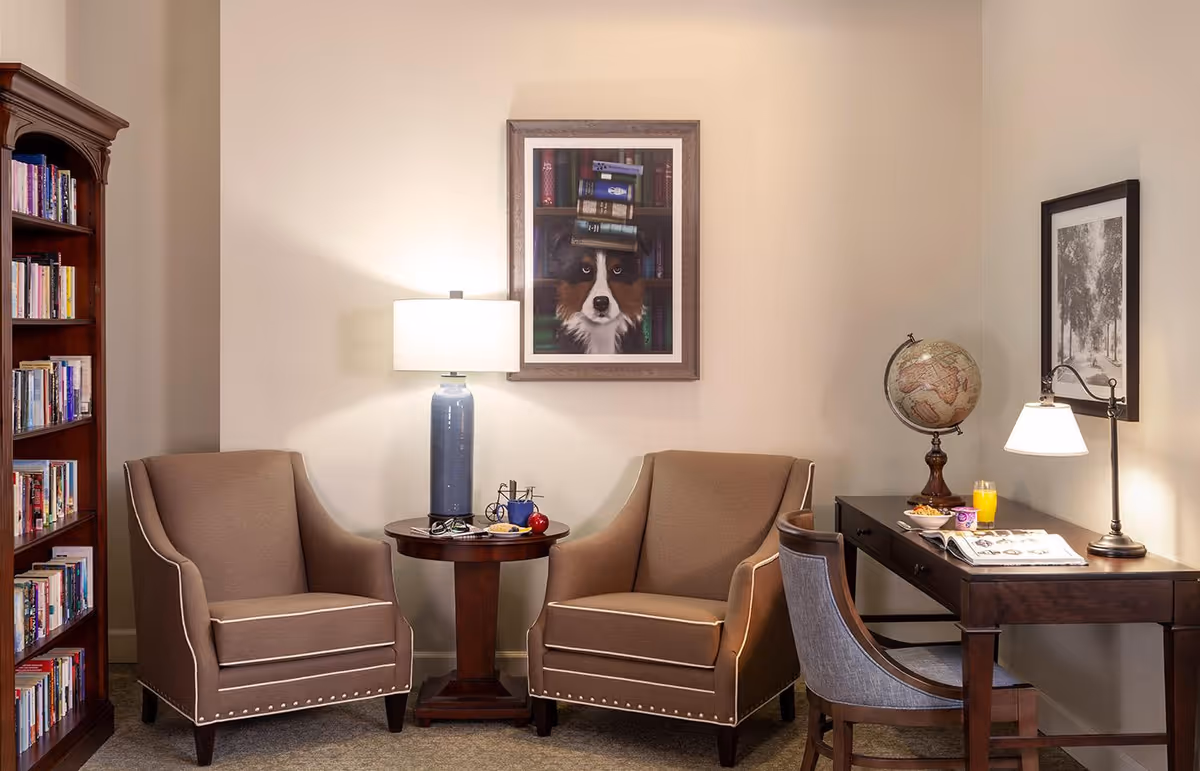 A cozy sitting area with two beige armchairs separated by a round wooden side table with a blue lamp, glasses, and an apple. To the right, there is a wooden desk with a globe, a lamp, a glass of orange juice, and an open book. A wooden bookshelf filled with books is on the left side of the image. The walls are decorated with framed pictures, including one of a dog with books on its head.
