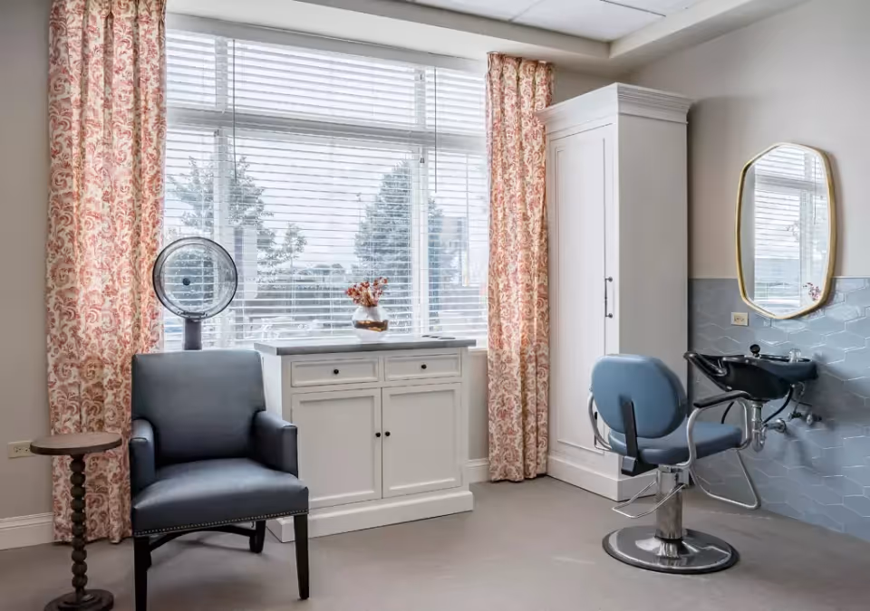 A bright room with large windows covered by white blinds and floral curtains. The room features a blue salon chair in front of a black wash basin with a gold-framed mirror above it. There is a white cabinet with drawers and a vase of flowers on top, a blue upholstered armchair, a small round wooden side table, and a hair dryer hood near the window.