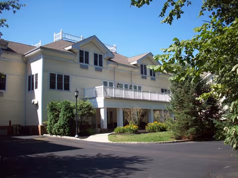 Exterior view of a two-story assisted living facility building with white siding, multiple windows, a balcony, and surrounded by trees and shrubs under a clear blue sky.