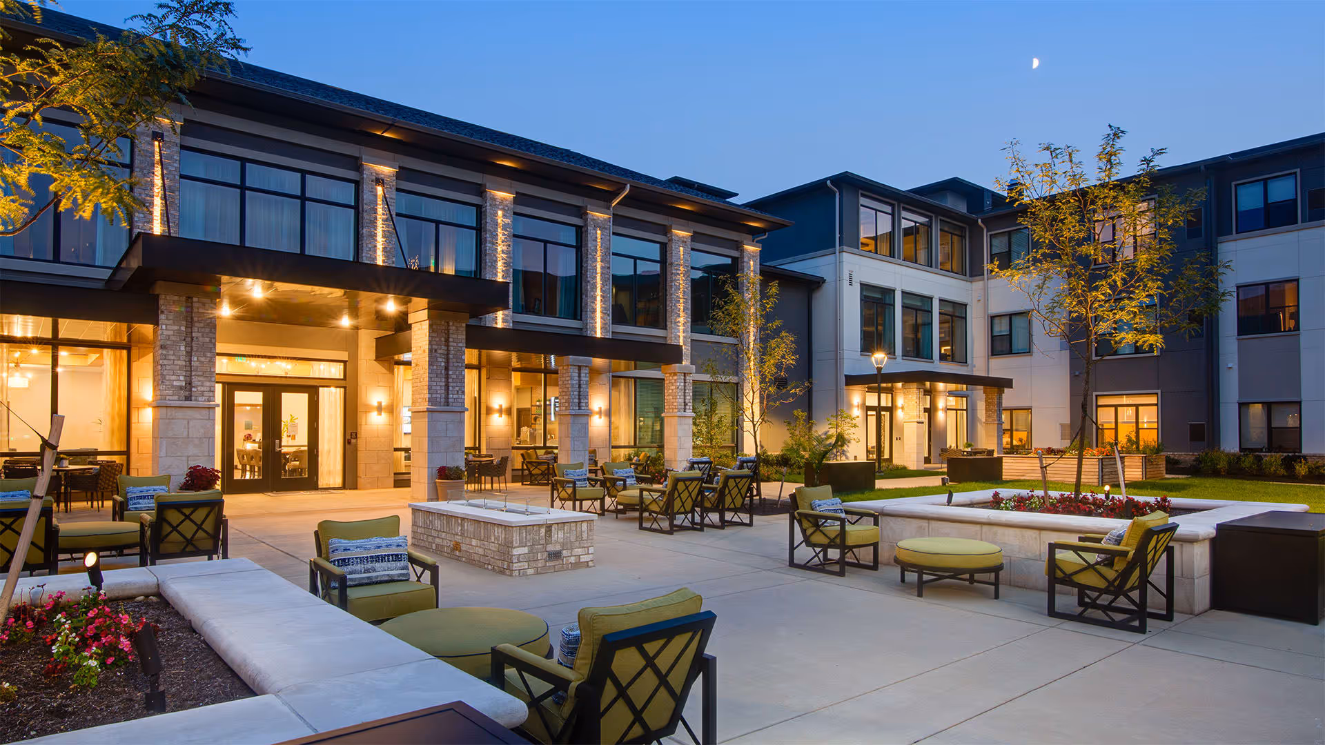 Outdoor patio area of Ridgecrest at Cranberry Woods senior living facility at dusk, featuring modern seating with green cushions, flower beds, trees, and a well-lit building exterior with large windows.