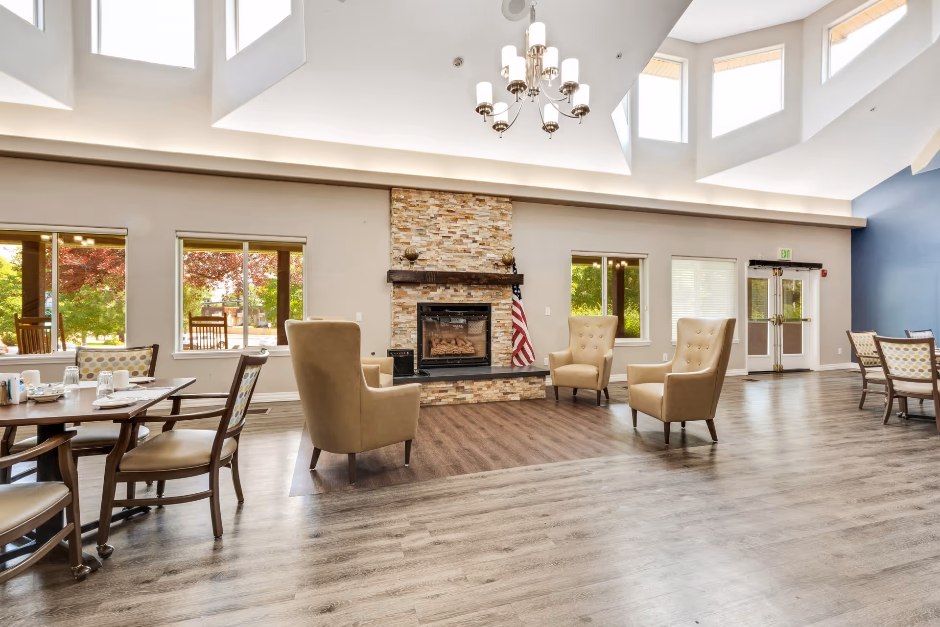 Spacious senior living common room with dining tables, armchairs around a stone fireplace and large skylights.