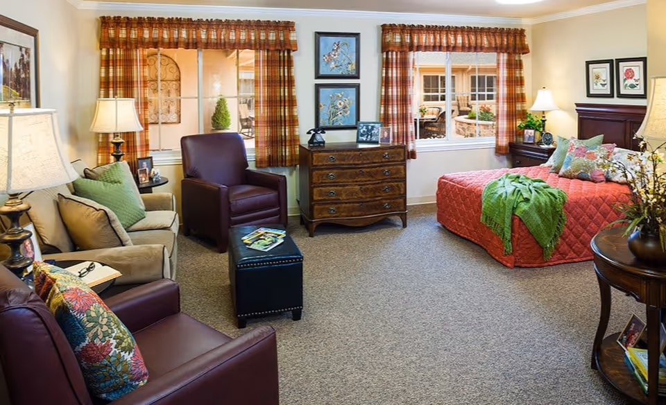 A cozy and well-decorated bedroom in a senior living facility featuring a bed with a red quilt and green throw, two windows with plaid curtains, a wooden dresser with decorative items, two armchairs with colorful cushions, a small ottoman, side tables with lamps, and framed artwork on the walls.