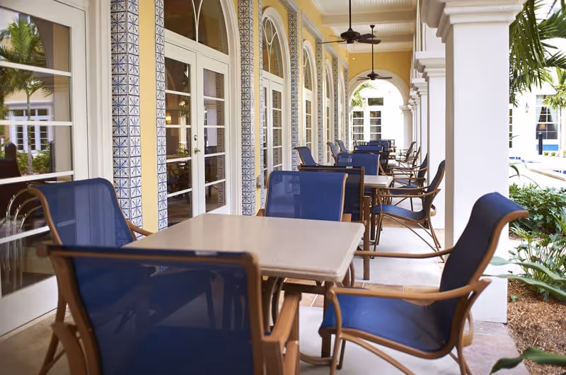 Covered outdoor patio dining area with rows of tables and blue chairs beside arched windows and columns.