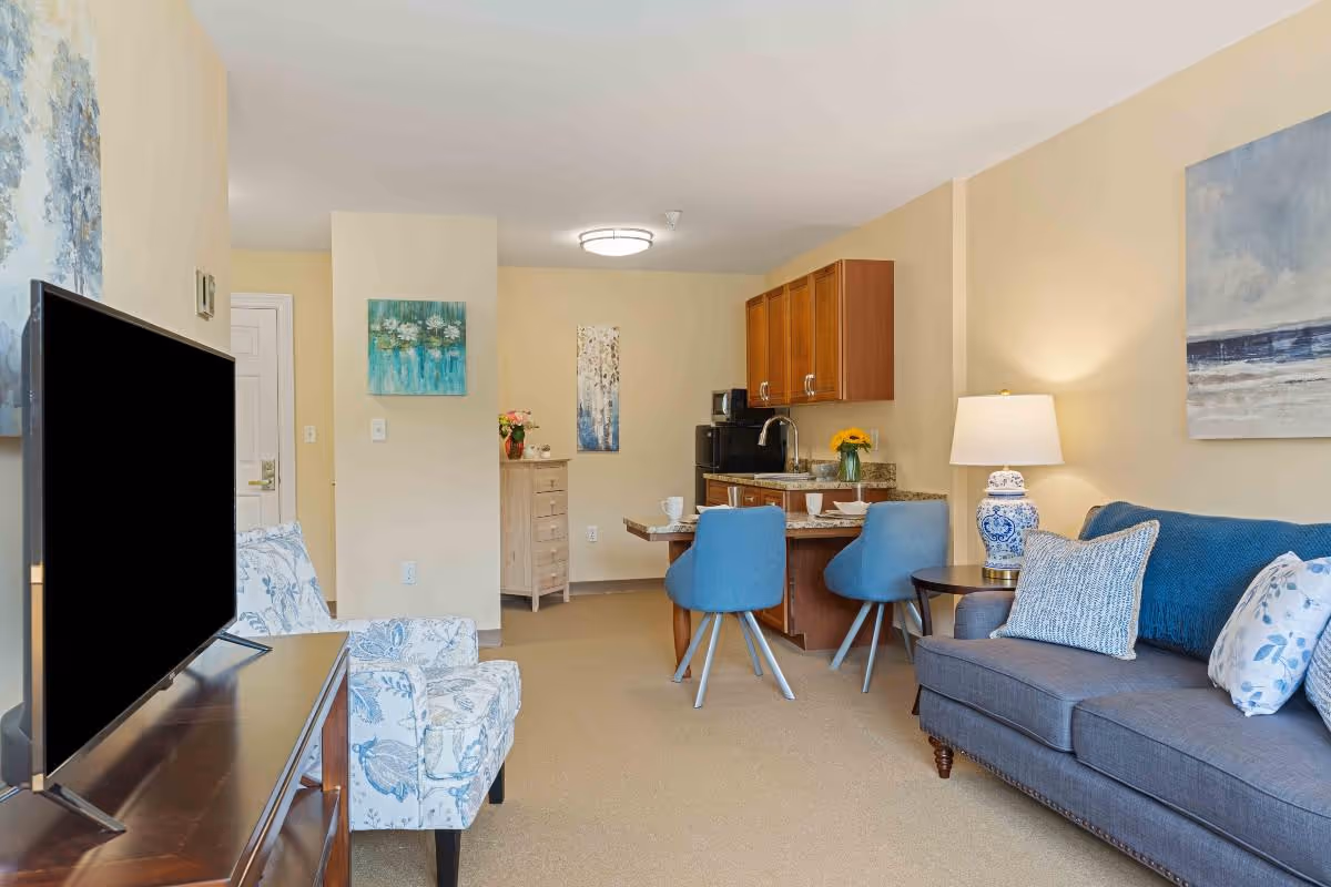 A cozy living area in a senior living facility featuring a gray sofa with blue and white pillows, a blue patterned armchair, a wooden TV stand with a flat-screen TV, and a small dining area with two blue chairs and a wooden table. The background includes a kitchenette with wooden cabinets, a countertop, and a small dresser with decorative items. The walls are painted light beige and decorated with paintings.