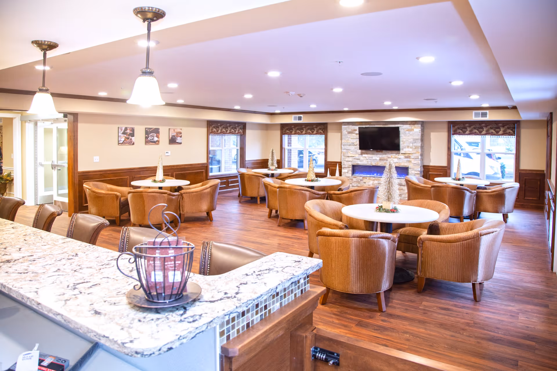 A spacious and well-lit common area with multiple round tables surrounded by comfortable brown upholstered chairs. The room features wooden flooring, a stone fireplace with a mounted flat-screen TV above it, and large windows with decorative valances. There is a granite countertop in the foreground with a decorative metal basket on it. The walls are painted beige with wooden wainscoting, and the ceiling has recessed lighting and hanging pendant lights.