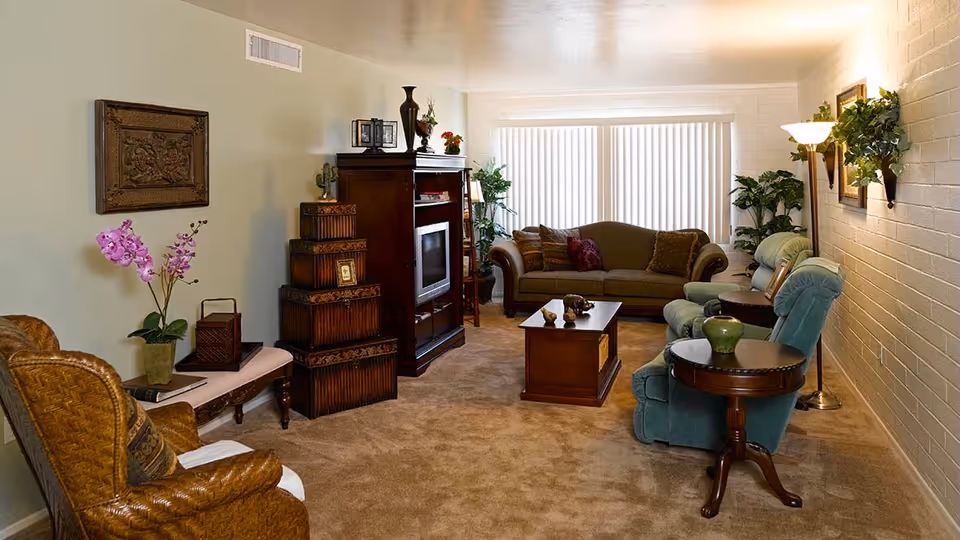 A cozy living room with beige carpet, a green sofa, two green armchairs, a wooden coffee table, and a wooden entertainment center with a TV. There are decorative plants, a floor lamp, and framed artwork on the walls. The room is softly lit with natural light coming through vertical blinds on a large window.