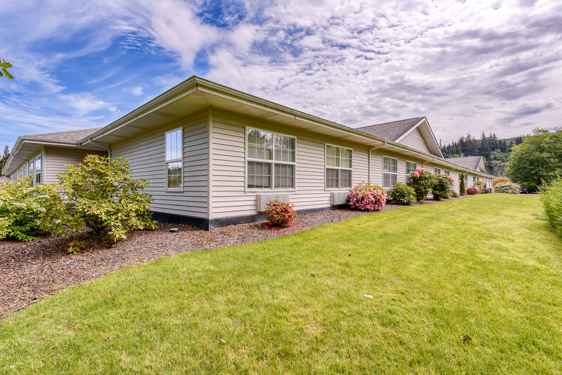 Single-story beige memory care building with large windows, landscaped shrubs and a green lawn under a partly cloudy sky.