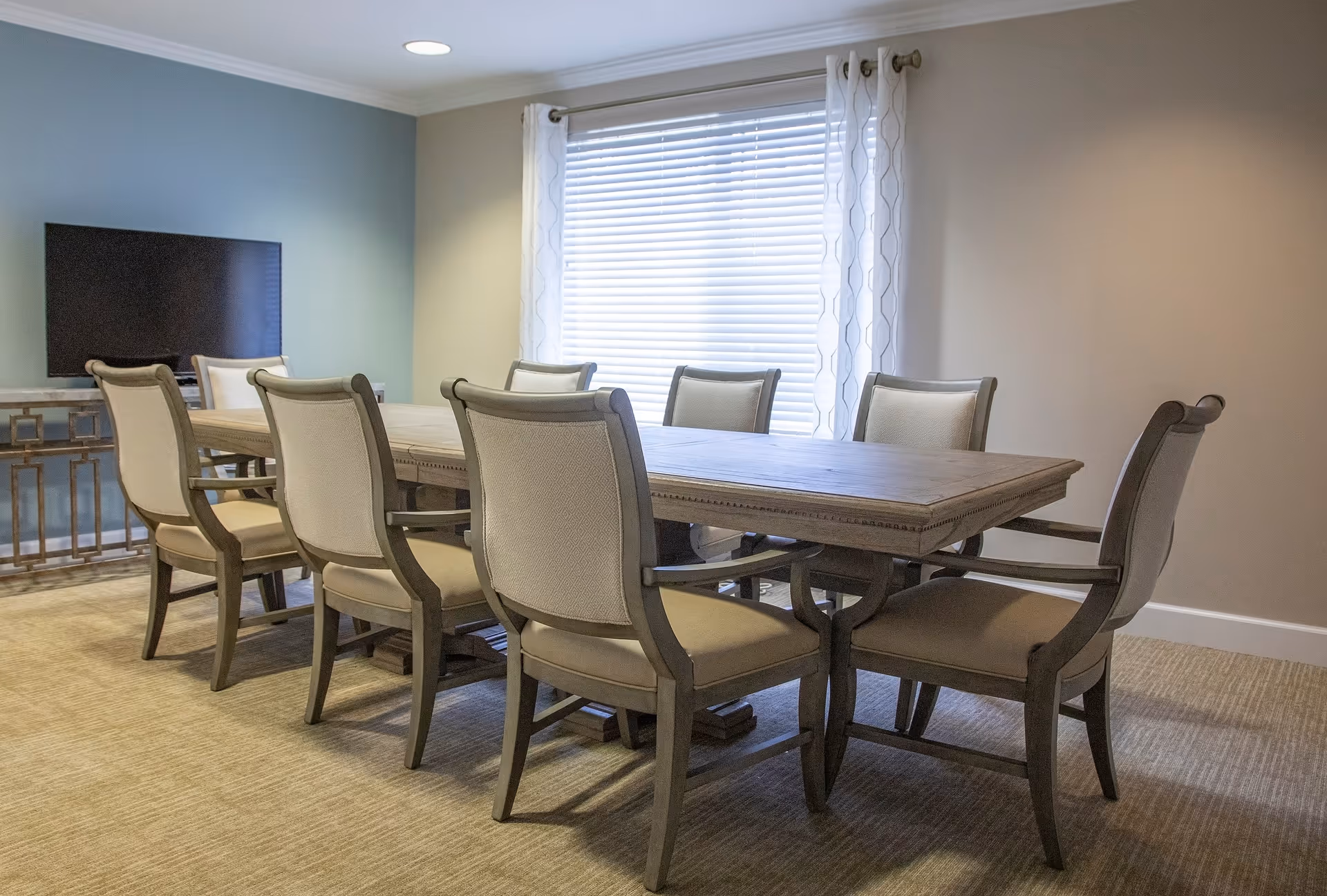A dining room with a long wooden table surrounded by eight upholstered chairs. A large window with white blinds and sheer curtains lets in natural light. A flat-screen TV is mounted on a blue accent wall with a decorative console table beneath it. The room has beige walls and carpeted flooring.