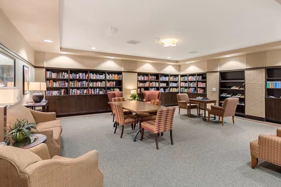 Spacious library-style common room with wall bookshelves, a central wooden table surrounded by upholstered chairs, and several armchairs.