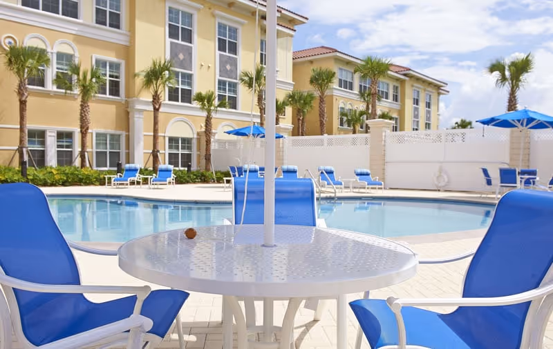 Outdoor swimming pool and patio with blue lounge chairs and umbrellas in front of a yellow, three-story building.