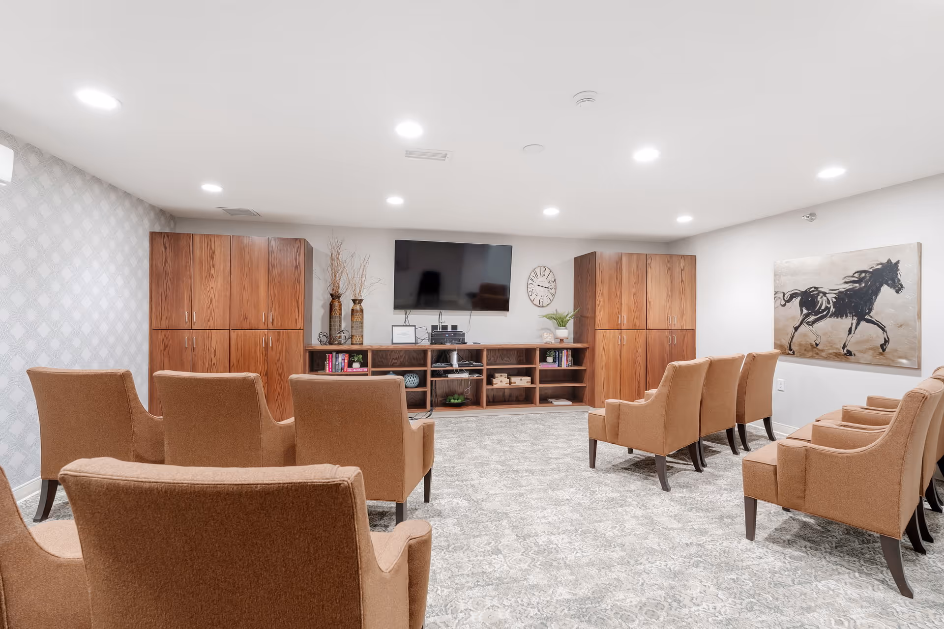 A bright, modern common room with beige armchairs arranged in rows facing a wall-mounted flat-screen TV. The room features wooden cabinets on either side of the TV, decorative vases, a clock, and a painting of a black horse on the right wall. The floor is carpeted with a patterned design, and the ceiling has recessed lighting.