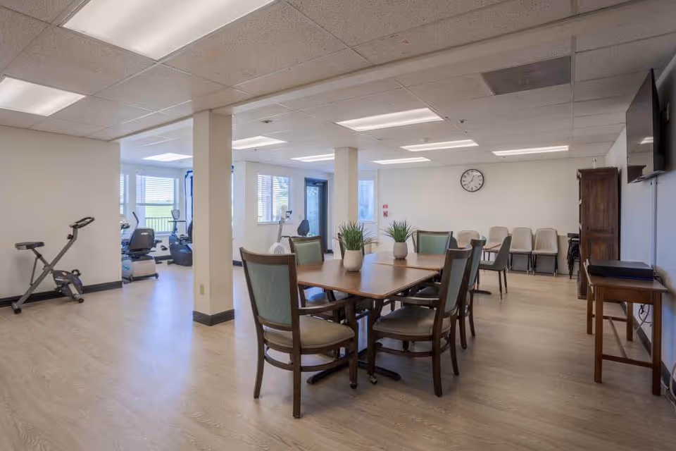 A spacious room with a wooden table and six chairs arranged around it, decorated with three small potted plants. In the background, there are several exercise machines near windows letting in natural light. The room has light-colored walls and ceiling with fluorescent lighting, a wall clock, and additional chairs lined up against the far wall. A TV is mounted on the right wall above a small wooden table.