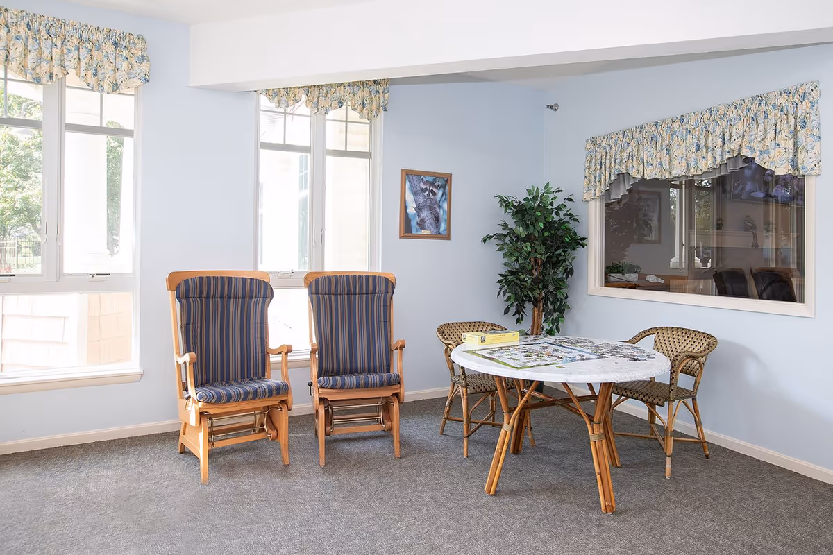 A small sitting area in a senior living facility with two wooden rocking chairs with blue striped cushions, a round table with a puzzle in progress, two wicker chairs, a potted plant, and windows with floral valances.