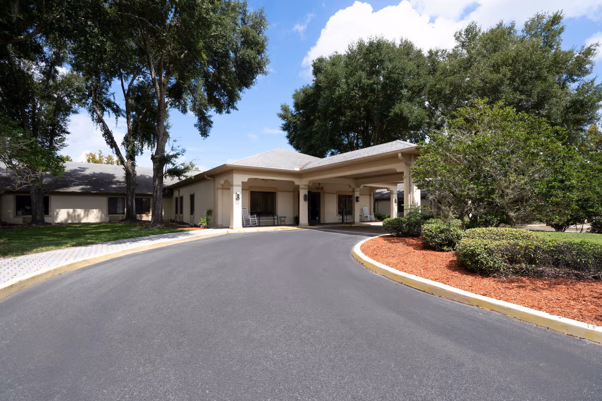 Driveway leading to the covered front entrance of a single-story senior living facility with trees and landscaped beds.
