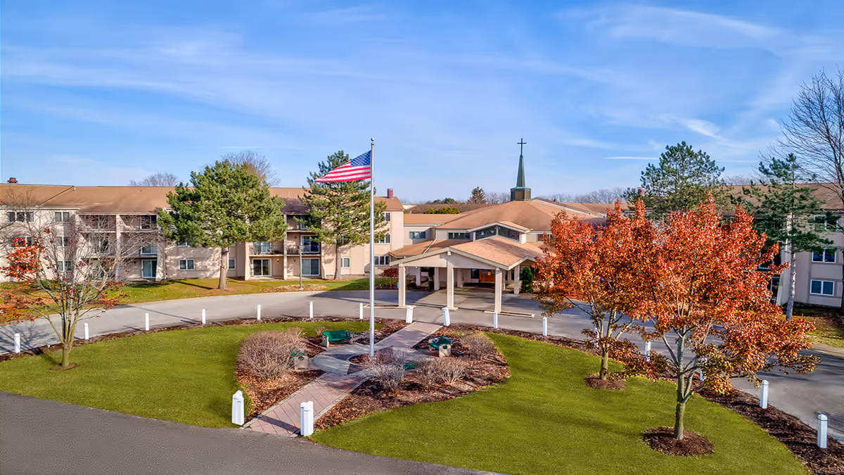 Exterior view of Winter Village senior living facility showing a large building with multiple windows and a covered entrance. There is a circular driveway with a flagpole flying the American flag in the center, surrounded by green grass, trees with autumn foliage, and landscaped bushes under a clear blue sky.