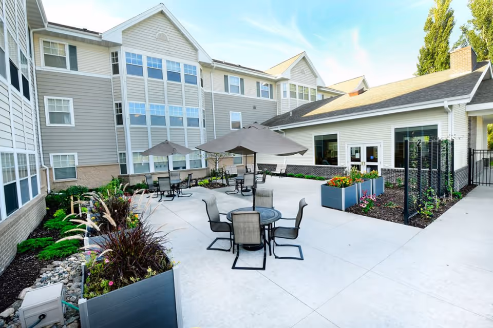 Outdoor courtyard at a senior living facility with patio tables and umbrellas, raised planters, and the surrounding multi-story building.