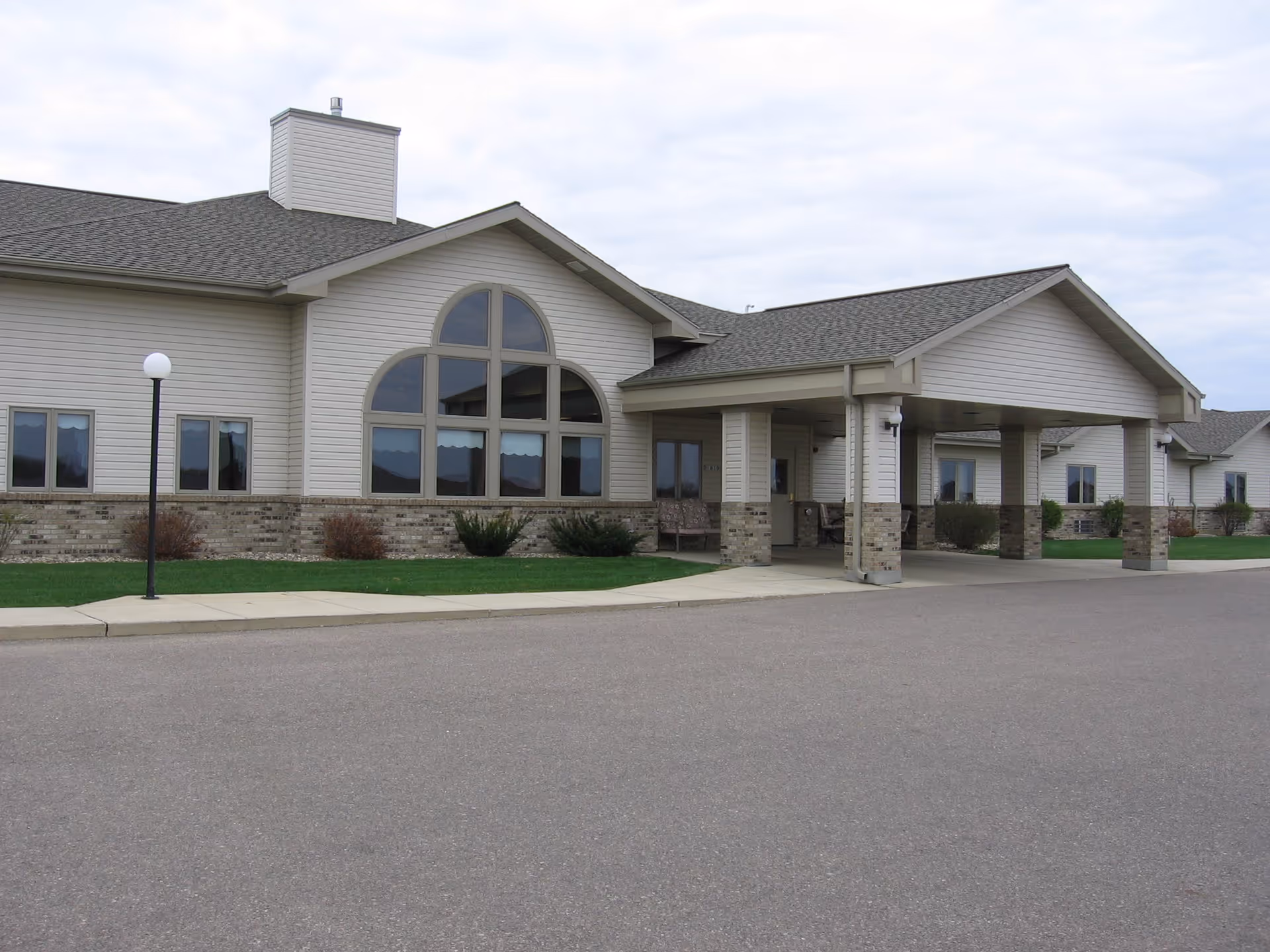 Exterior view of a single-story building with beige siding and brick accents, featuring large arched windows and a covered entrance with columns. The building is surrounded by a paved driveway, green grass, and a lamppost under a cloudy sky.