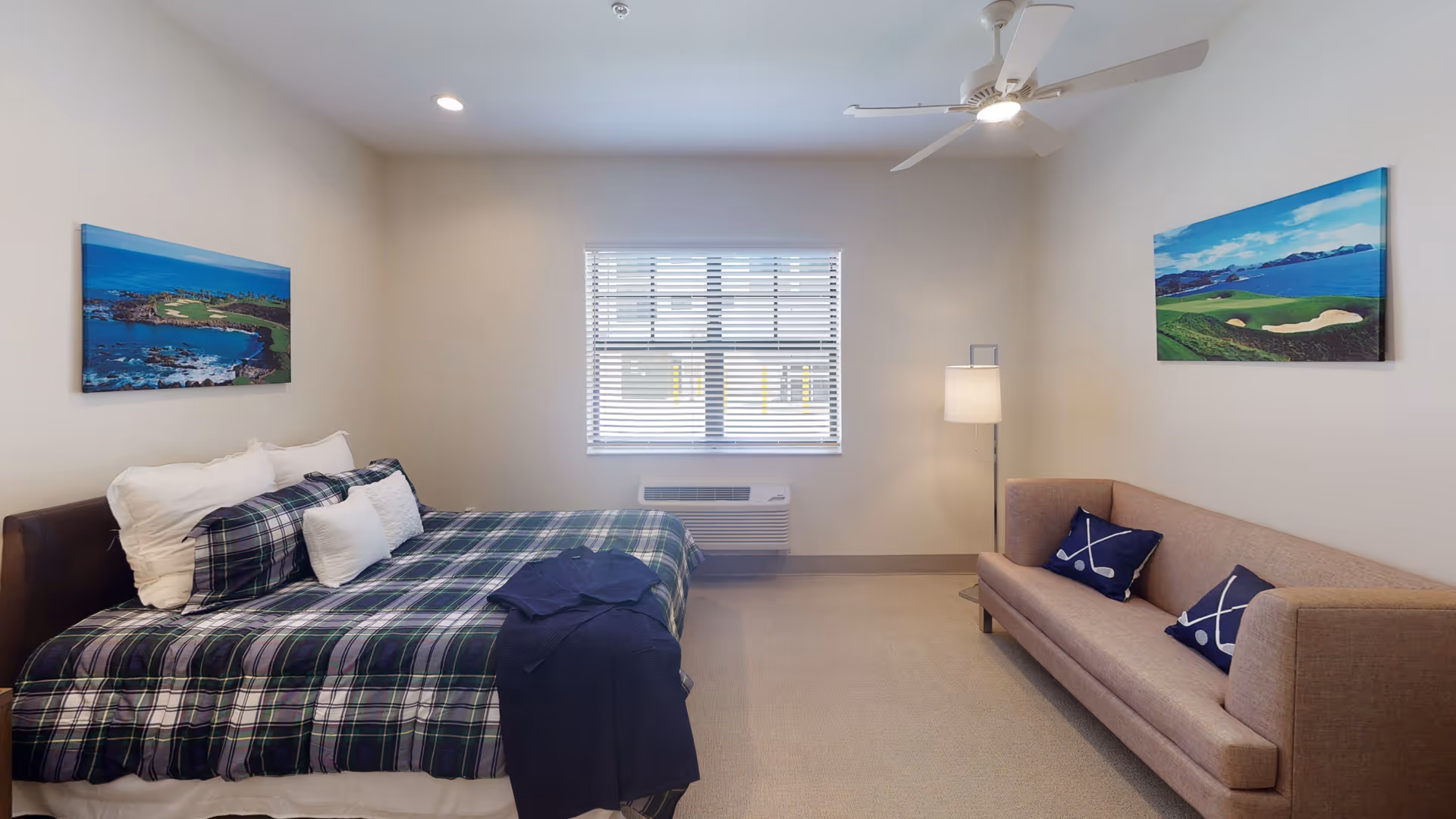 A neatly arranged bedroom with a bed on the left side covered with a plaid blanket and multiple pillows. There is a beige sofa on the right side with two decorative pillows featuring crossed golf clubs and golf balls. The room has a window with blinds in the center, a floor lamp next to the sofa, and two landscape paintings of golf courses on the walls. A ceiling fan is mounted above.