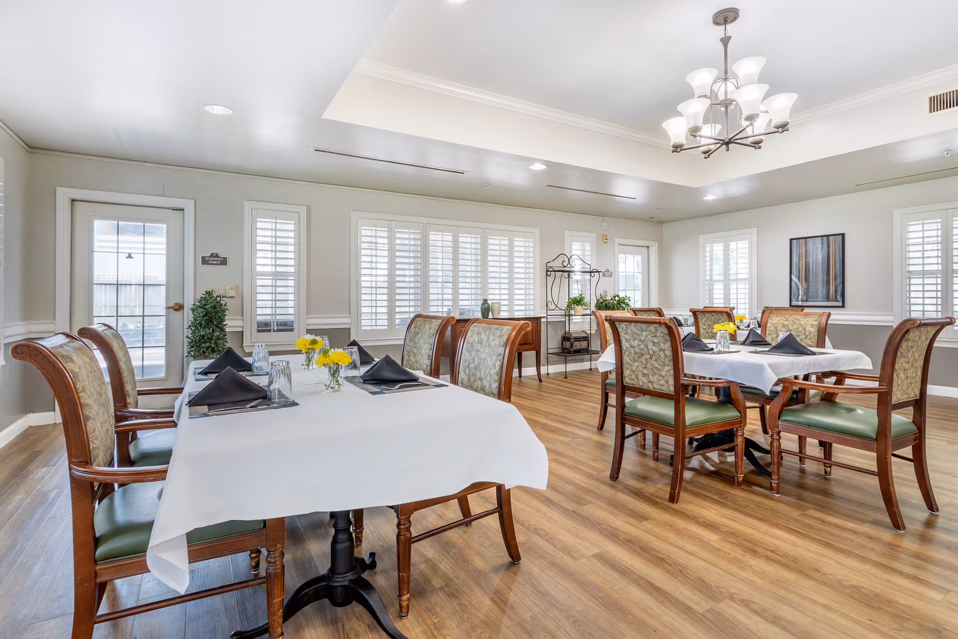 A bright dining room with wooden floors and multiple tables covered with white tablecloths. Each table is set with black napkins, glasses, and small vases of yellow flowers. The room has large windows with white shutters, a chandelier hanging from the ceiling, and wooden chairs with patterned upholstery.