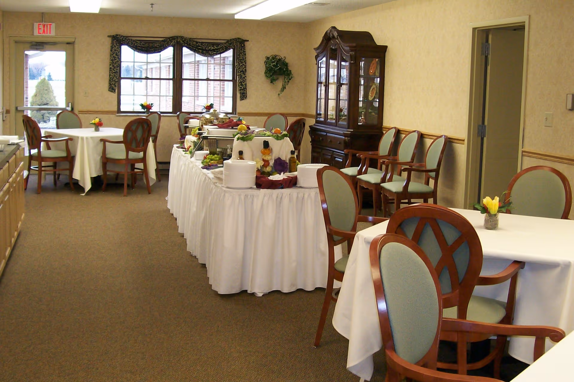 A dining room with several round tables covered with white tablecloths and surrounded by wooden chairs with green cushions. In the center of the room, there is a long buffet table draped with a white skirt, displaying various food items and decorations. A wooden china cabinet is against the wall, and a window with a valance lets in natural light. An exit door is visible in the background.