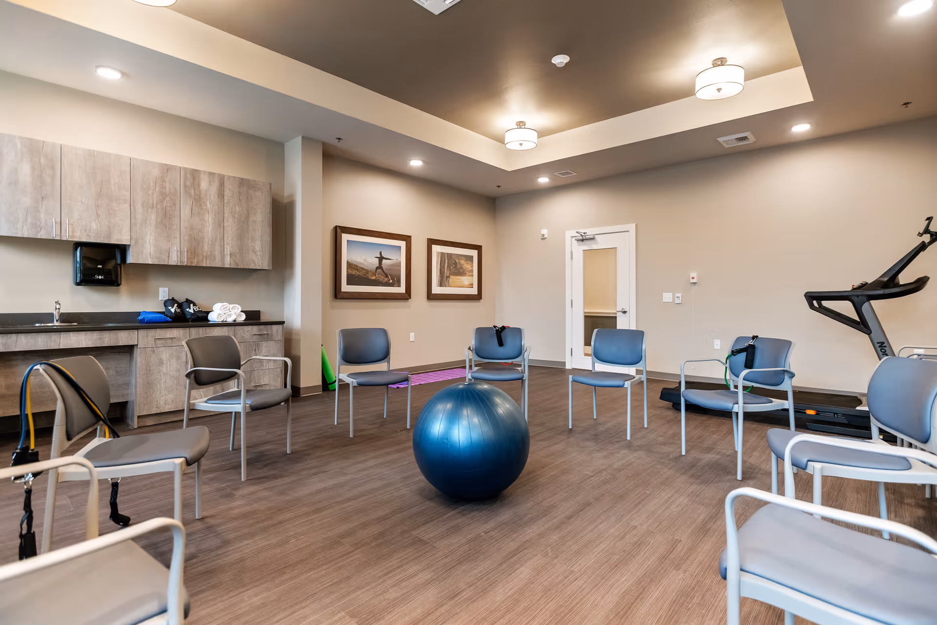 A spacious exercise room with several chairs arranged in a circle around a large blue exercise ball. The room has wood flooring, beige walls, and ceiling lights. There is a treadmill and exercise equipment in the corner, a countertop with cabinets, and two framed pictures on the wall.