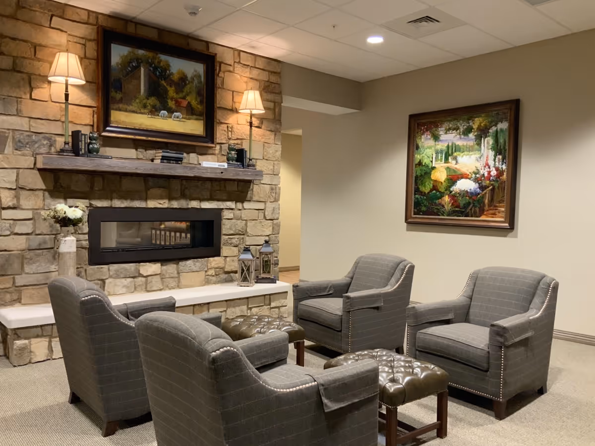 A cozy sitting area with four gray upholstered armchairs arranged around two tufted leather ottomans in front of a stone fireplace. The fireplace has a wooden mantel with two lamps and decorative items. Above the mantel is a framed painting of a countryside scene with sheep. Another colorful framed painting of a garden is hanging on the adjacent wall.
