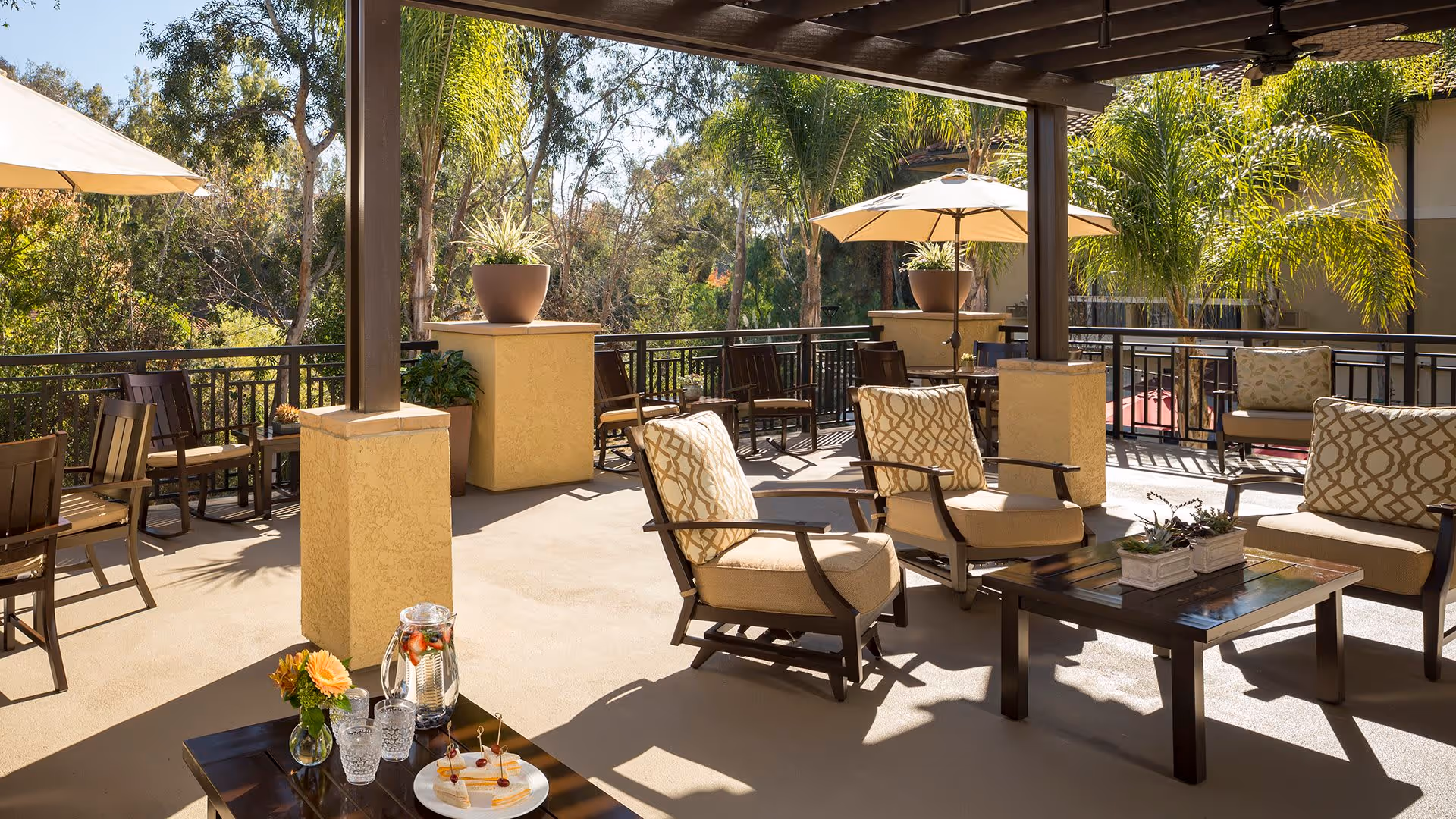 Outdoor patio area with cushioned chairs and wooden tables under a pergola. There are potted plants, umbrellas providing shade, and a table with a pitcher of infused water, glasses, and a plate of appetizers. Trees and greenery are visible in the background.