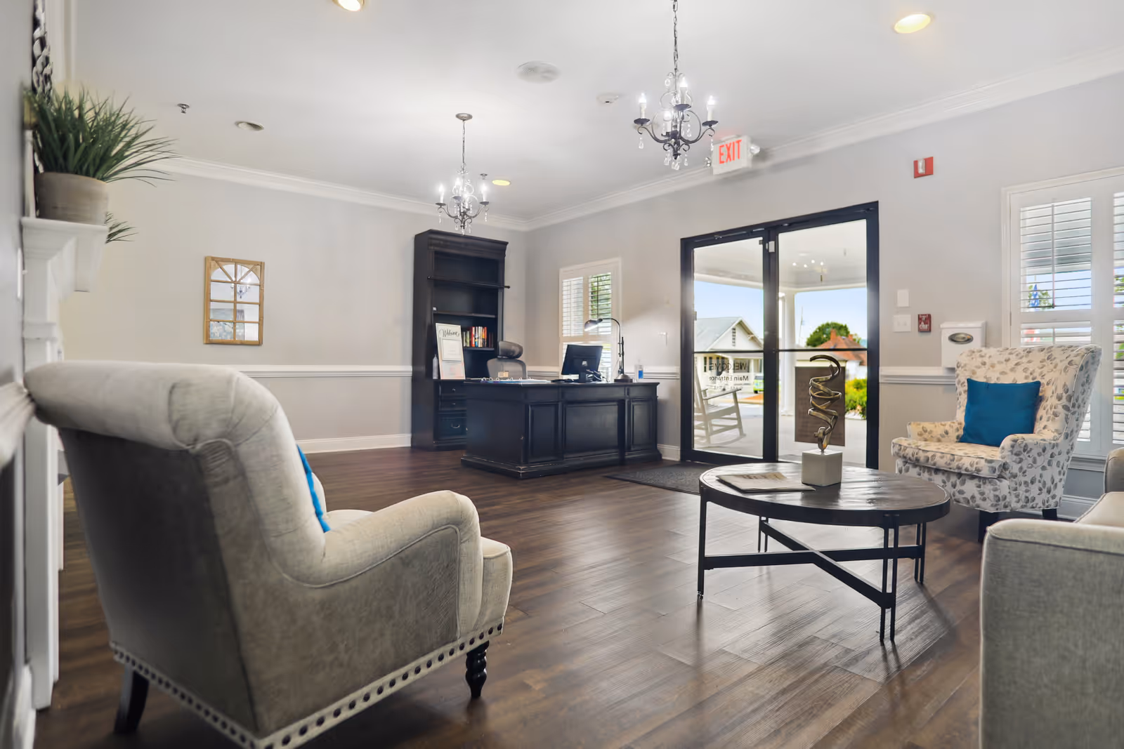 A bright and welcoming reception area with a dark wooden desk, a computer, and a bookshelf behind it. The room features comfortable upholstered chairs with blue accent pillows, a round coffee table with a decorative sculpture, and large glass doors leading outside. The floor is wooden, and the walls are painted light gray with white trim. Two chandeliers hang from the ceiling, and there are windows with white shutters allowing natural light to enter.