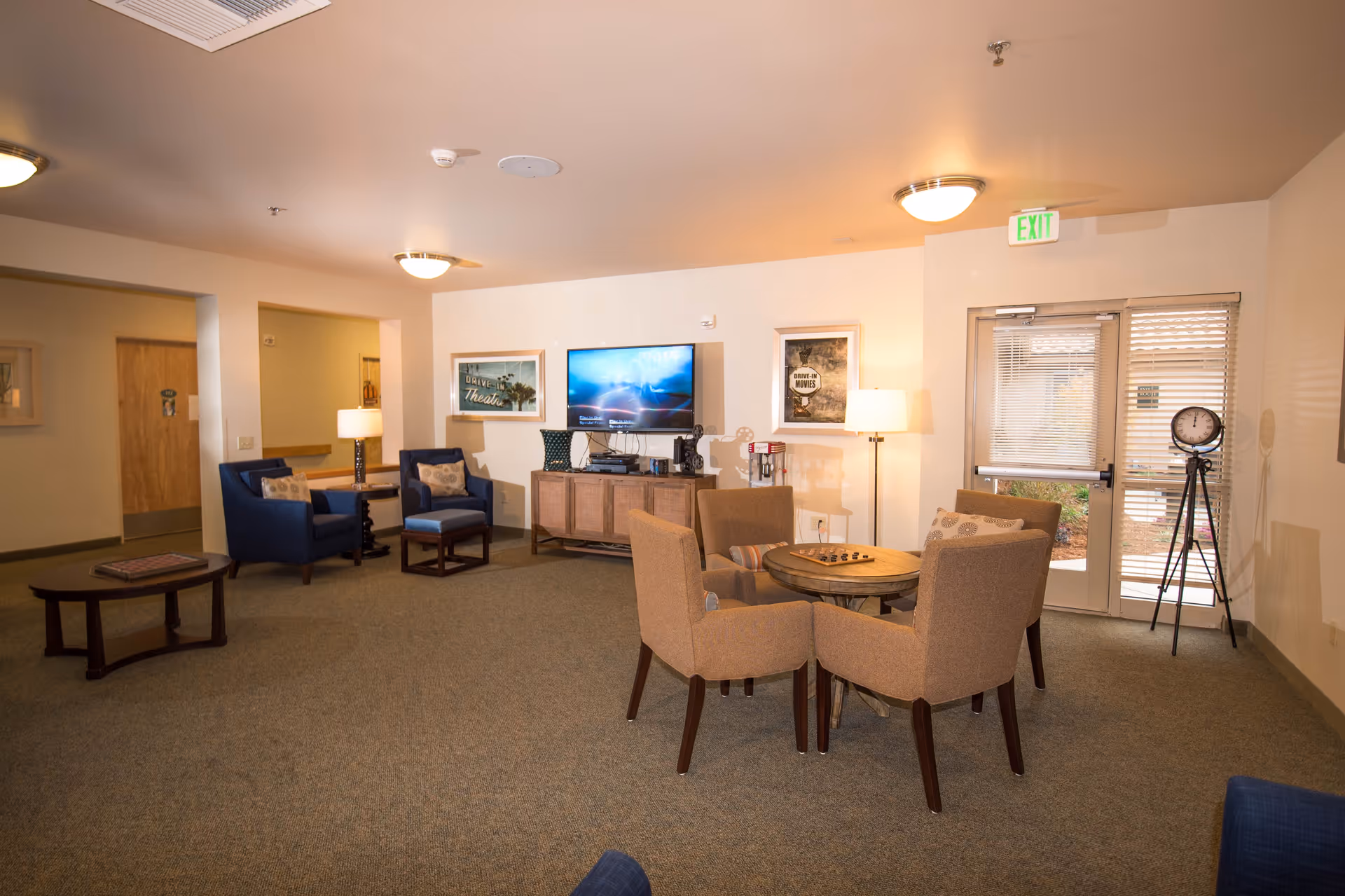 A cozy living room area in a senior living facility with a round table and four beige chairs in the center. To the left, there are two blue armchairs with a small table and lamp between them. A flat-screen TV is mounted on the wall above a wooden cabinet. The room has carpeted floors, beige walls, ceiling lights, and a glass door leading outside. Decorative framed pictures hang on the walls, and a vintage-style clock on a tripod stands near the door.
