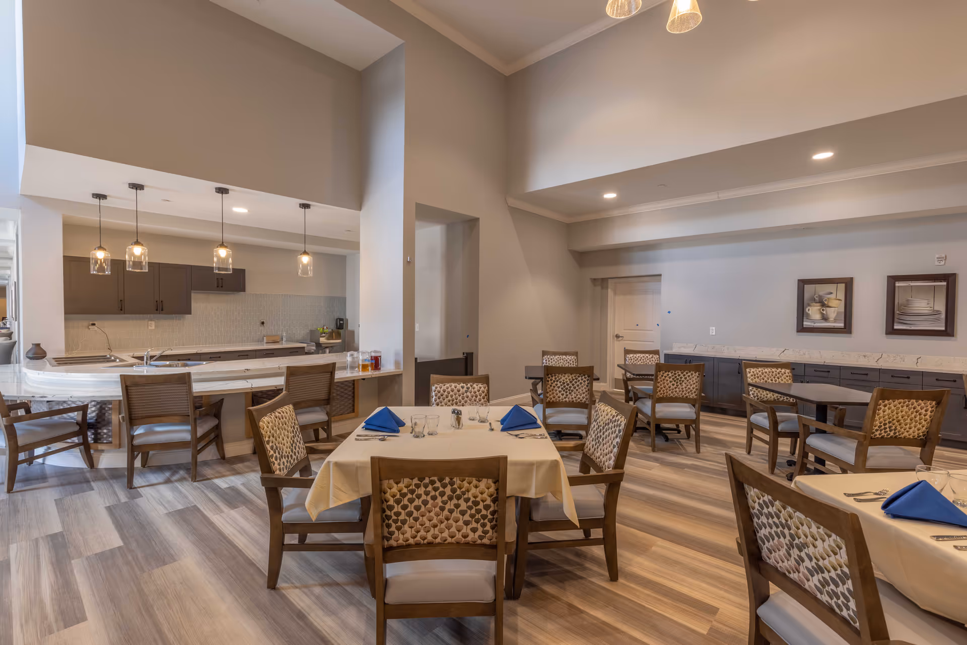 A spacious dining room with several tables covered with beige tablecloths and set with blue napkins, glasses, and silverware. The room features wooden chairs with patterned upholstery, a kitchen area with a countertop and hanging pendant lights, and light-colored wood flooring. The walls are painted in neutral tones with framed artwork of dishes hanging on one wall.