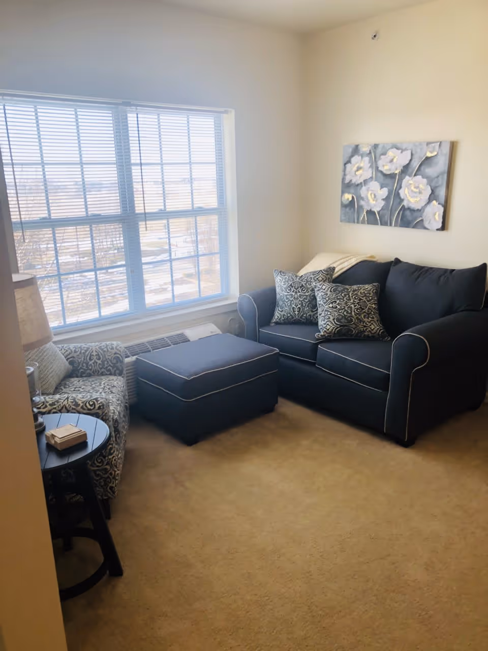 A cozy living room with a large window letting in natural light. The room features a dark blue loveseat with patterned throw pillows, a matching ottoman, a patterned armchair, a small round side table with a lamp and a book, and a floral painting on the wall.