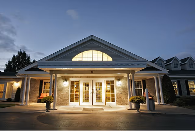 Front exterior view of a senior living facility building named Tennyson Court at dusk, featuring a well-lit entrance with double glass doors, white columns, and decorative plants on either side.