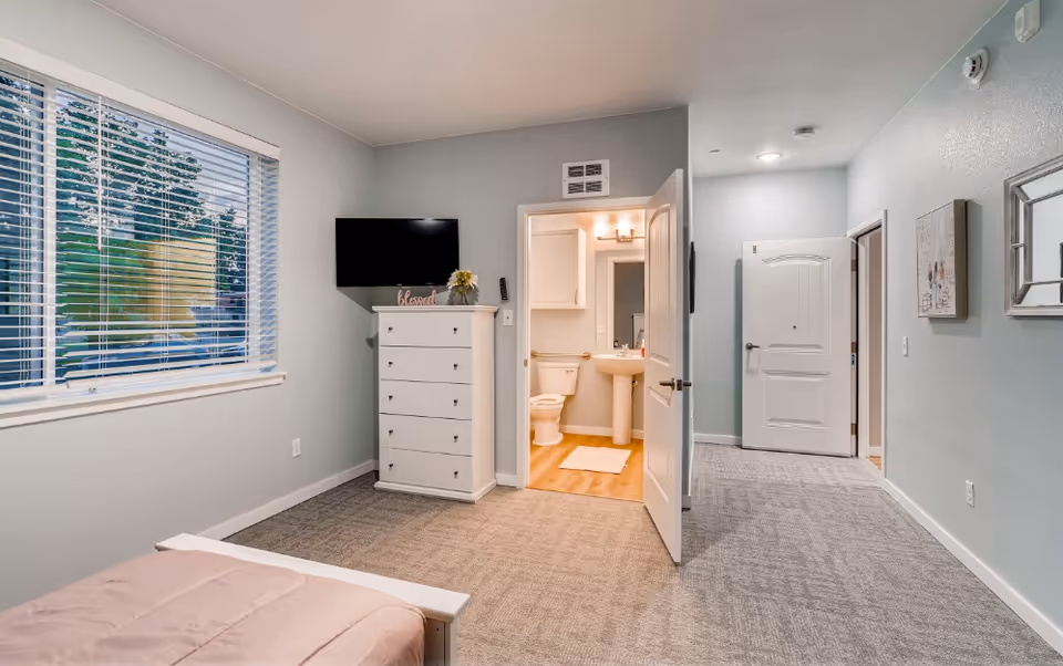Bright assisted-living bedroom with a bed in the foreground, a white dresser and wall-mounted TV, and an open door leading to an ensuite bathroom.