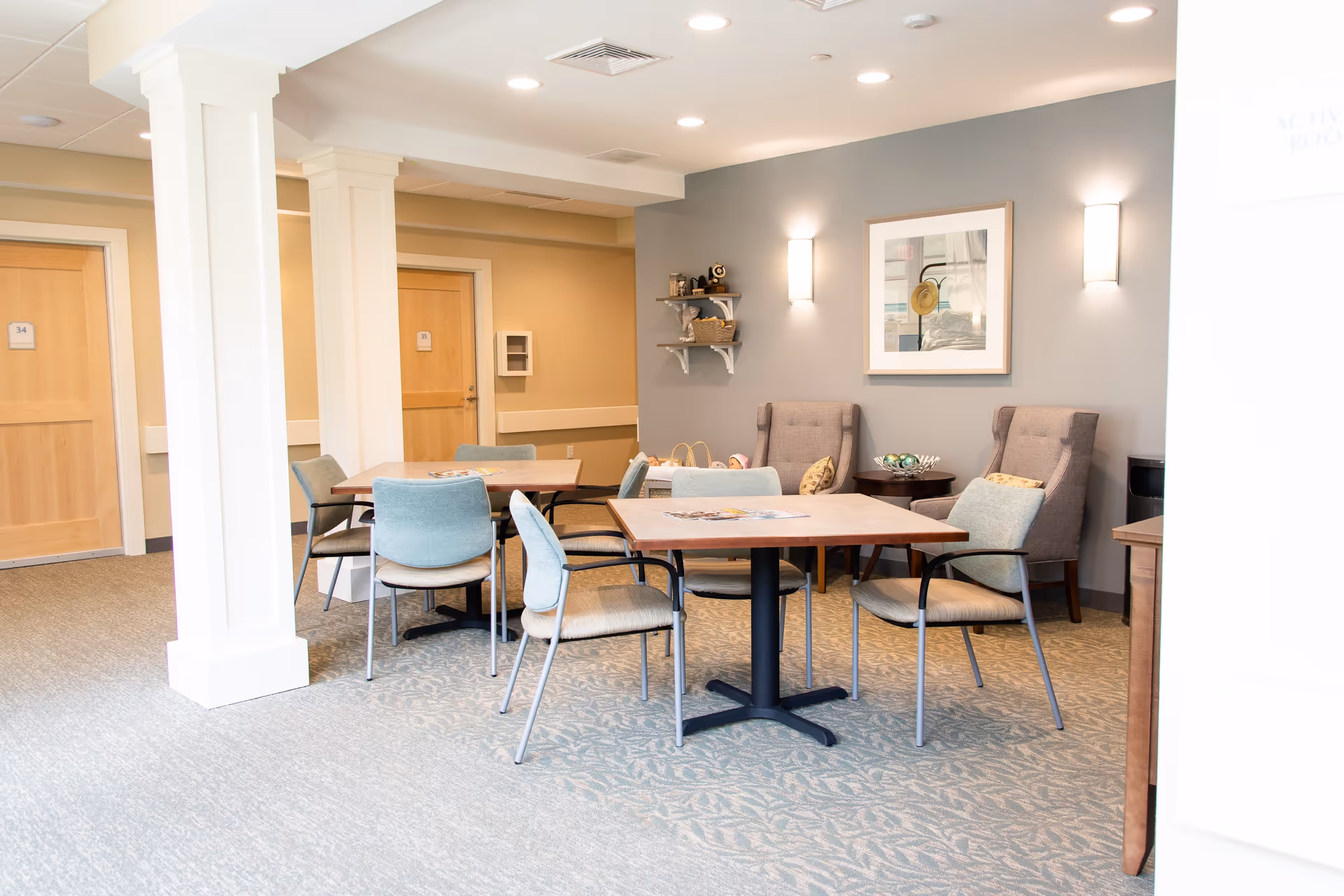 A well-lit room with two square tables surrounded by chairs, two upholstered armchairs with pillows, a small round side table, and wall-mounted shelves holding decorative items. The walls are painted in neutral tones with framed artwork and wall sconces providing additional lighting. Two wooden doors are visible in the background.