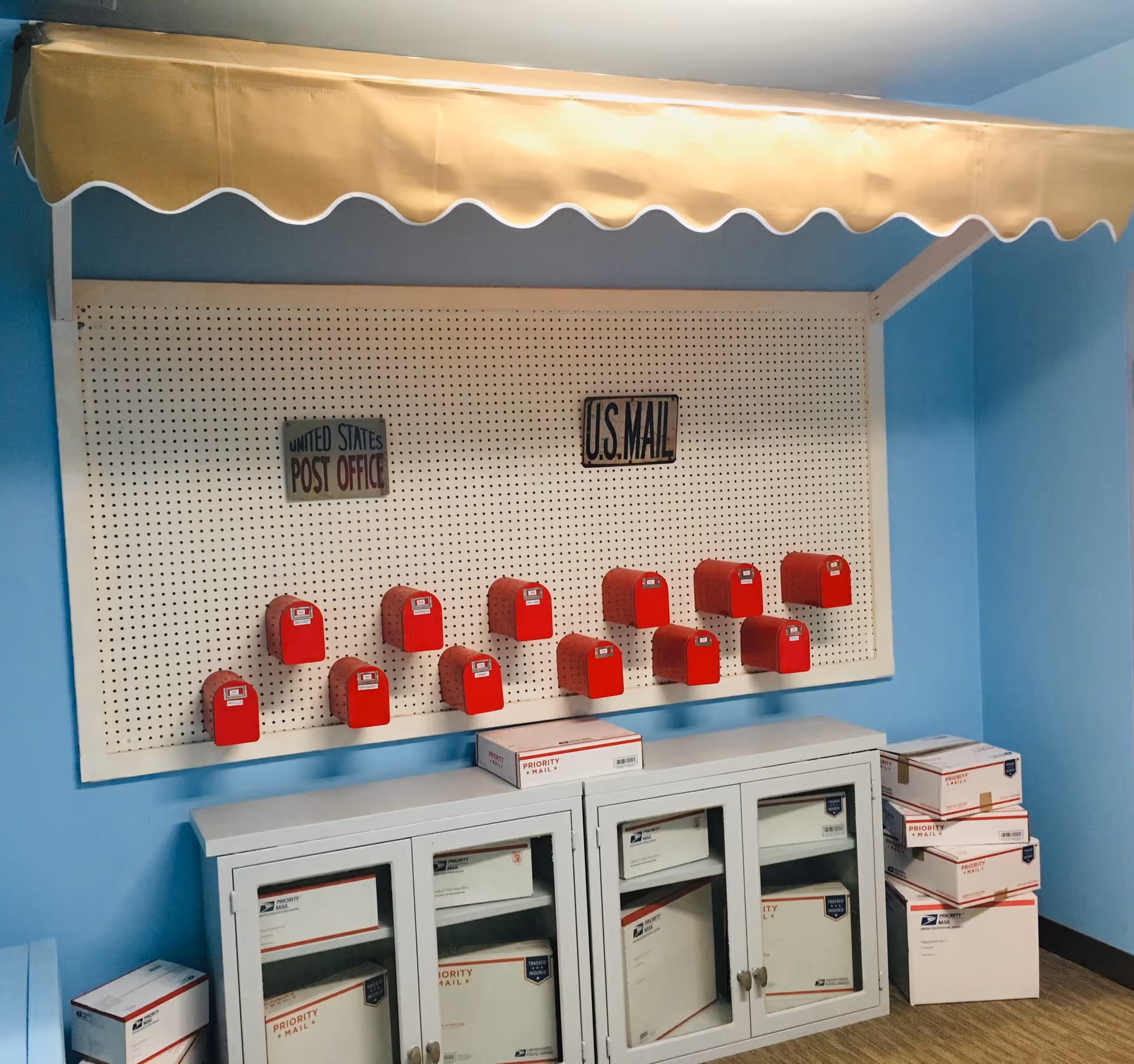 A mailroom area with a blue wall and a white pegboard displaying small red mailboxes. Above the mailboxes are two signs that read 'UNITED STATES POST OFFICE' and 'U.S. MAIL'. Below the pegboard is a white cabinet with glass doors containing Priority Mail boxes, and more Priority Mail boxes are stacked on the floor to the right and left of the cabinet. A yellow awning with a scalloped edge is mounted above the pegboard.