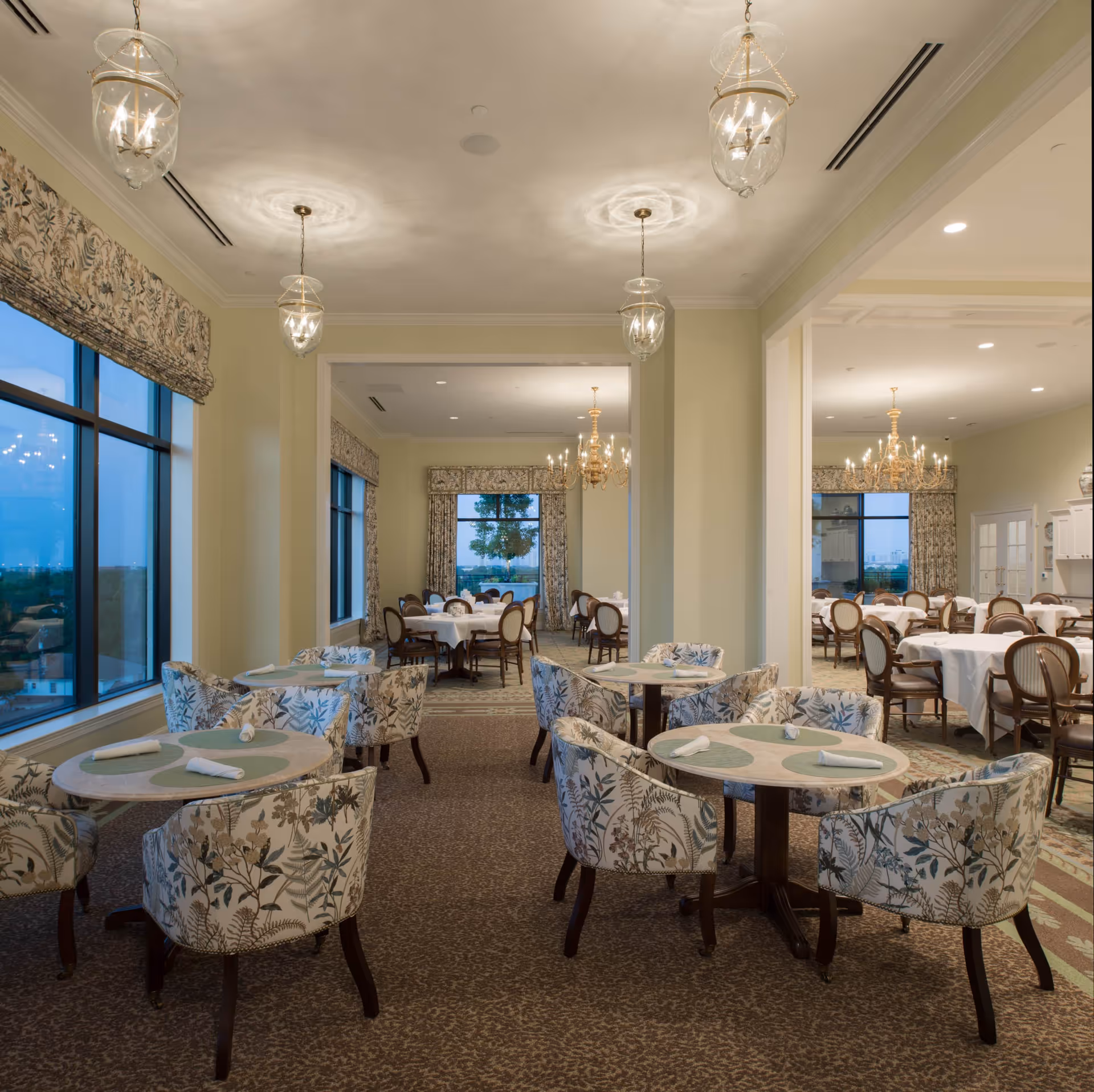 A spacious dining room with multiple round tables covered with white tablecloths and floral-patterned chairs. The room features large windows with patterned curtains, elegant chandeliers, and soft lighting creating a warm and inviting atmosphere.