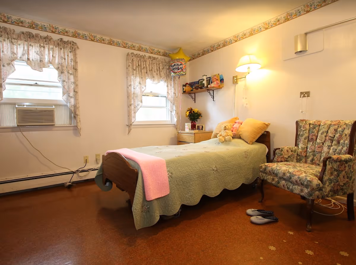 A cozy bedroom in a senior living facility with a single bed covered in a green quilt and a pink blanket at the foot. The bed has stuffed animals and pillows on it. Next to the bed is a floral upholstered armchair and a pair of slippers on the floor. Two windows with floral curtains and an air conditioning unit are on the left wall. A small wooden nightstand with flowers and a shelf with toys and decorations are on the far wall. A wall-mounted lamp provides warm lighting.