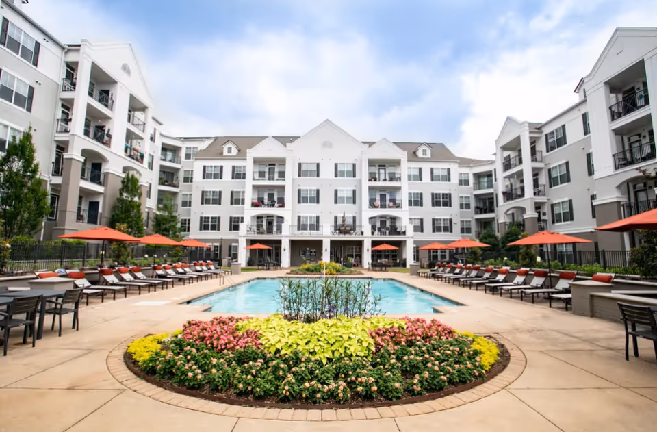 Outdoor swimming pool area at Emblem Alpharetta with lounge chairs and orange umbrellas surrounding the pool. The pool is centered with a circular flower bed featuring colorful plants in the foreground. White multi-story residential buildings with balconies surround the pool area under a partly cloudy sky.