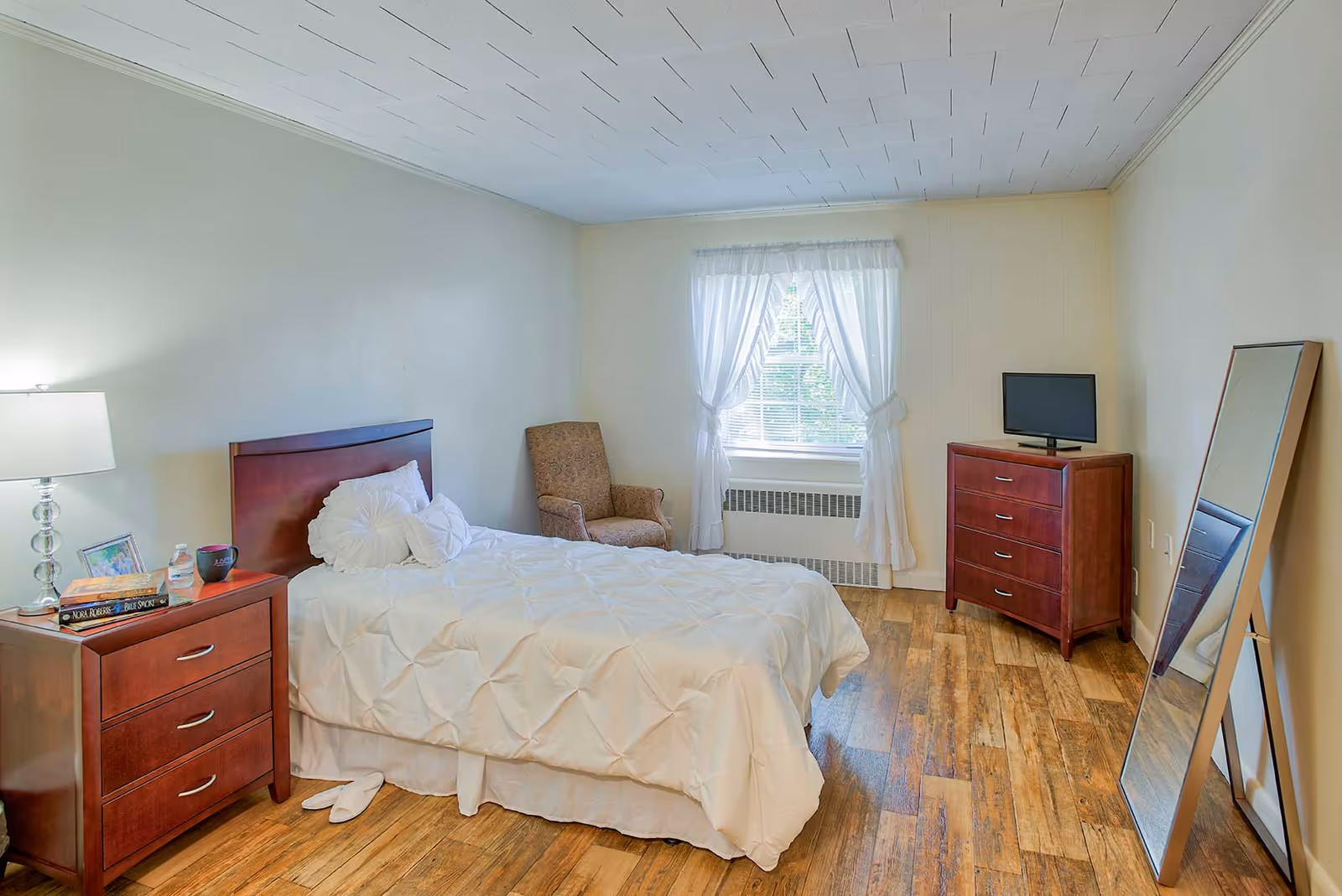 A cozy bedroom with a single bed covered in a white comforter and decorative pillows. Next to the bed is a wooden nightstand with a lamp, books, a framed photo, and a cup. There is a comfortable armchair near a window with white curtains letting in natural light. Across from the bed is a wooden dresser with a small TV on top and a large standing mirror leaning against the wall. The room has wooden flooring and light-colored walls.