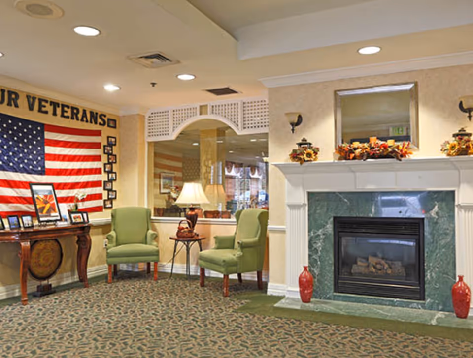 A cozy senior living facility common area with two green armchairs and a small table with a lamp between them. There is a fireplace with a green marble surround decorated with autumn-themed floral arrangements and candles. On the left wall, an American flag is displayed with the words 'OUR VETERANS' above it, along with framed photos and artwork on a wooden table beneath the flag. The room has patterned carpet and warm lighting.