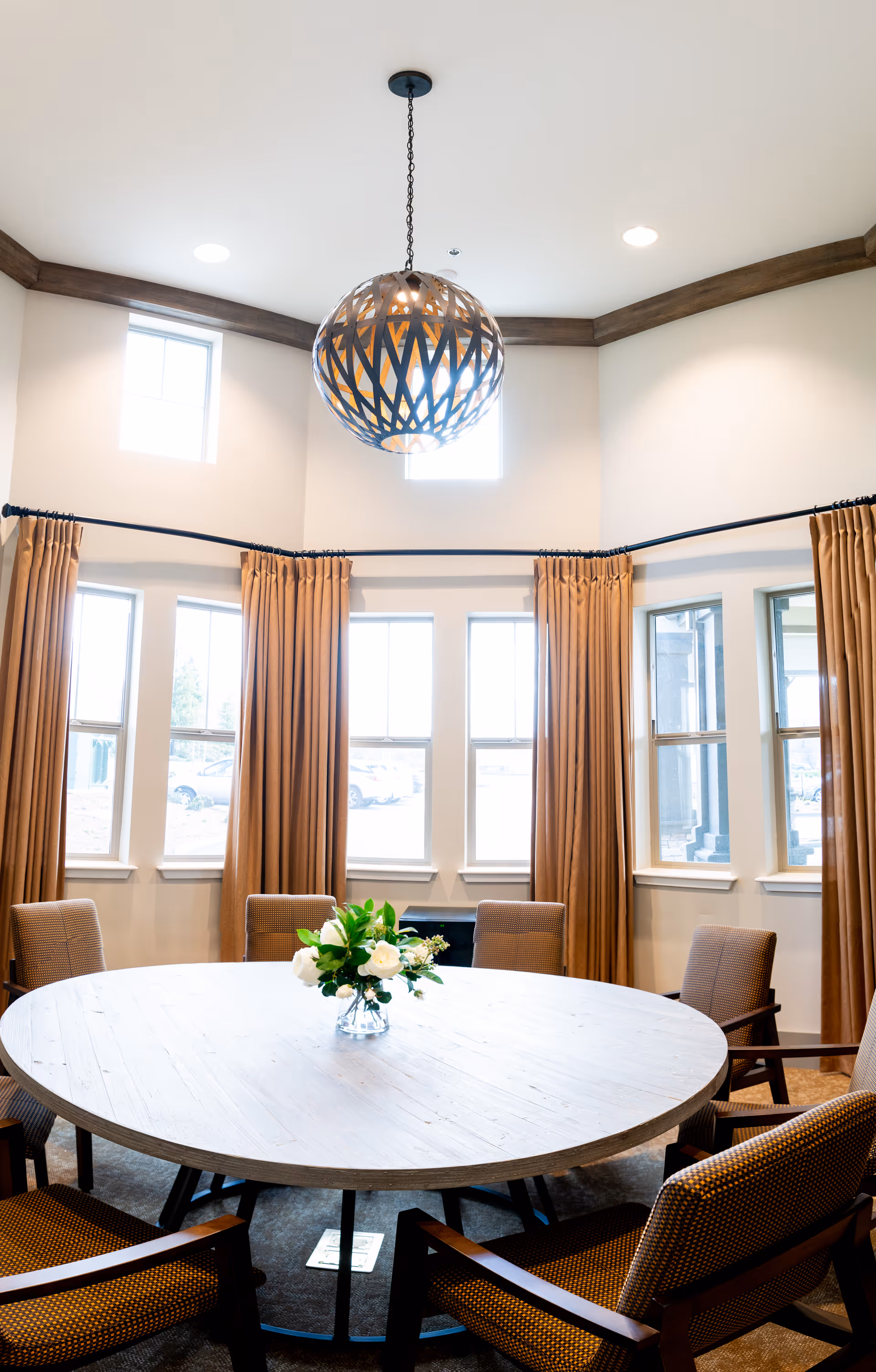 A bright room with a large round wooden table surrounded by six cushioned chairs. The room features multiple windows with brown curtains and a modern spherical pendant light hanging from the ceiling. A small vase with white flowers and greenery is placed in the center of the table.