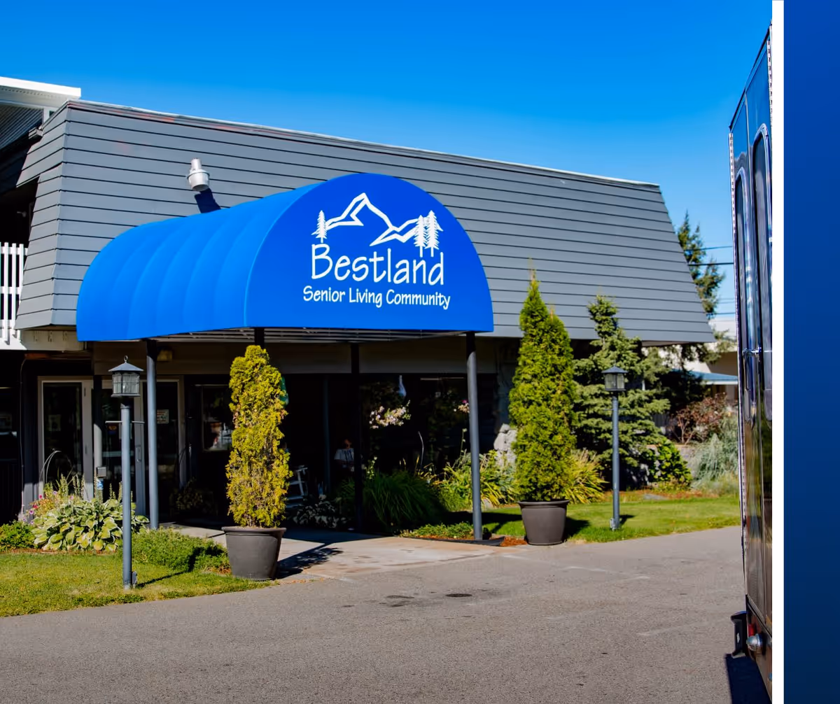 Front entrance of Bestland Senior Living Community with a blue awning, potted shrubs, and landscaping.